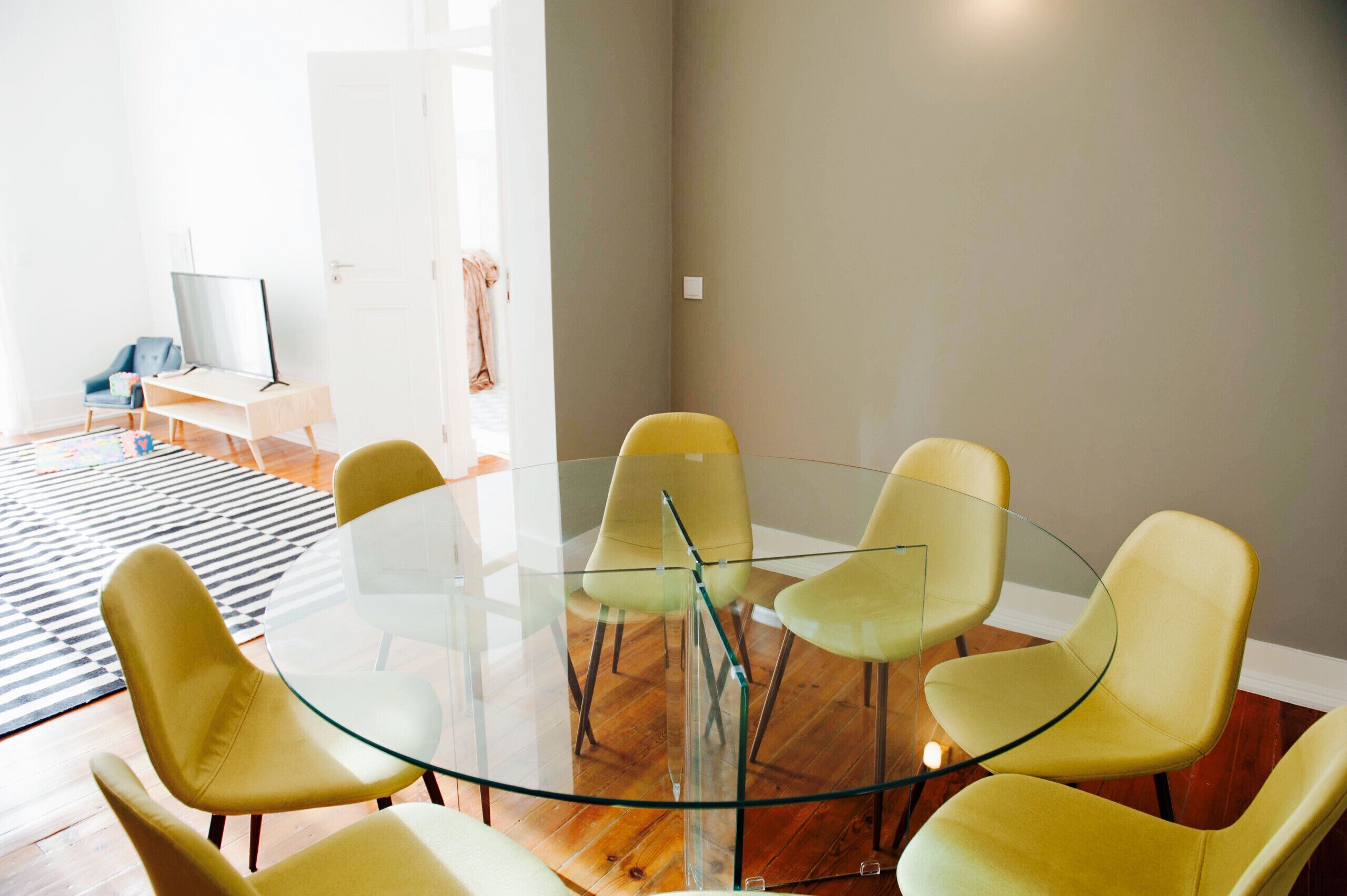Un salon moderne avec une table en verre entourée de chaises jaunes, un téléviseur posé sur un meuble et un tapis à rayures sur le sol.