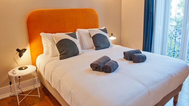 Una cama bien hecha con ropa blanca y dos toallas enrolladas, una lámpara de noche y una ventana con vistas a edificios.