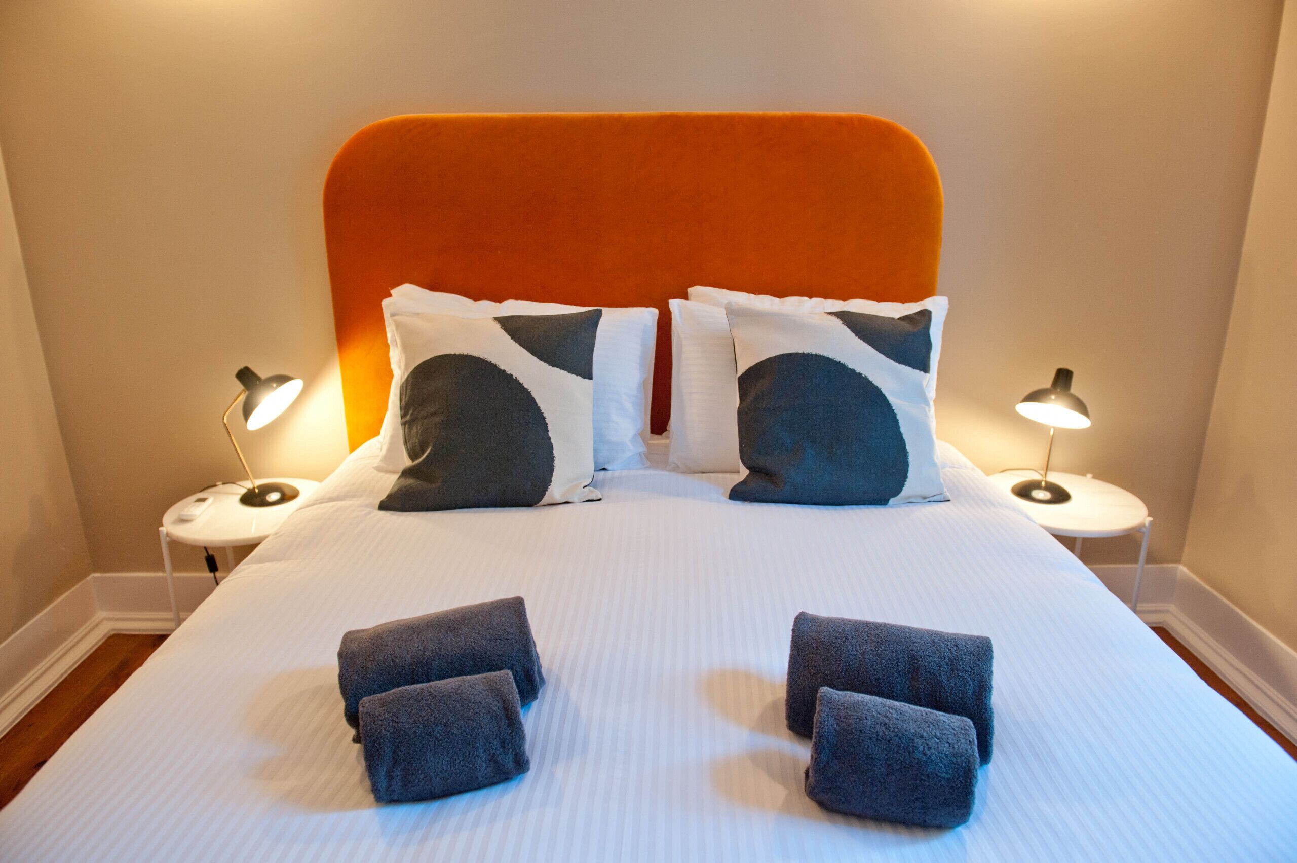 A neatly made bed with an orange headboard, two bedside lamps, and four towels on the bed.