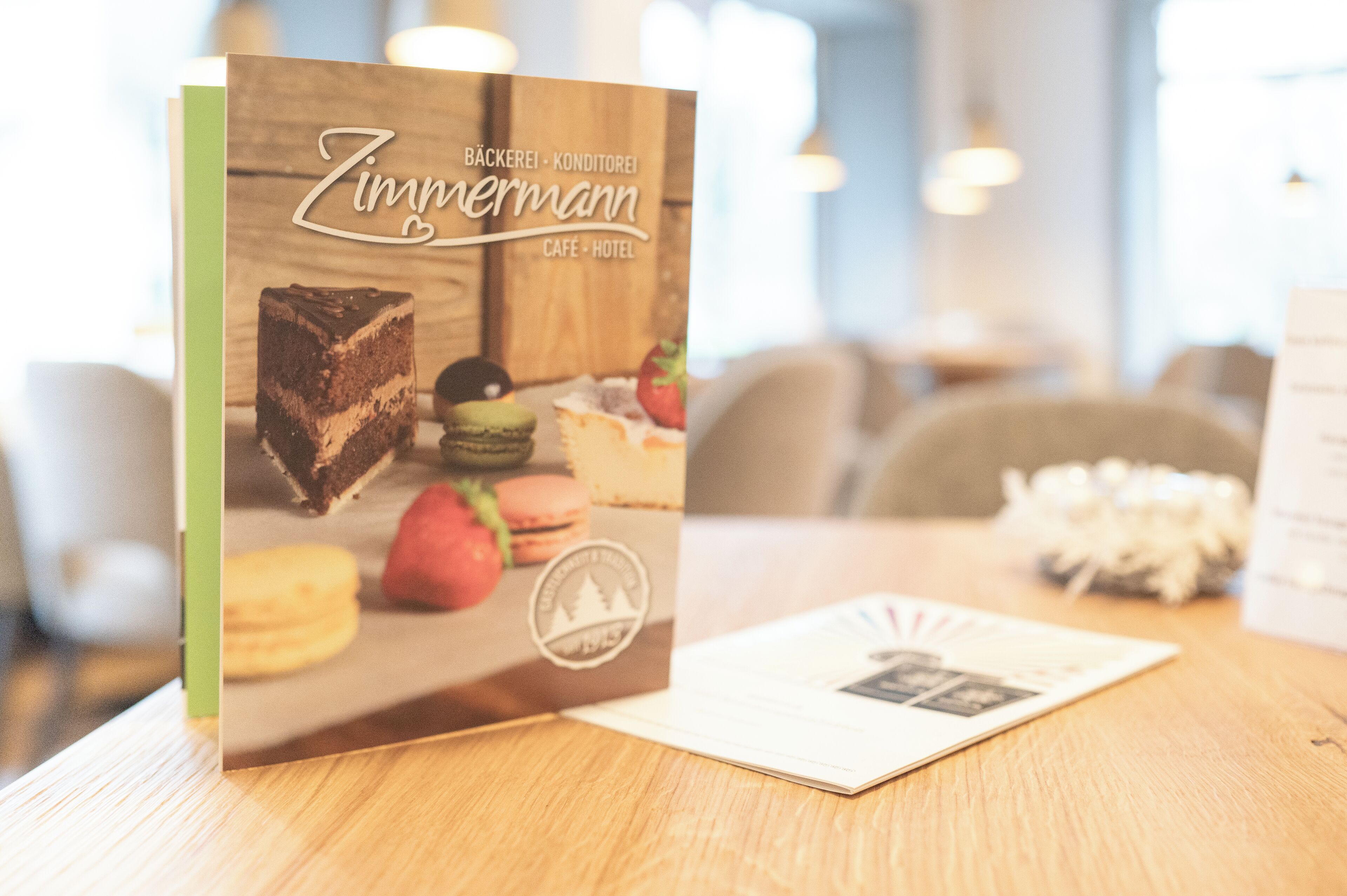 Un menu ou une brochure intitulée « Zimmermann », comportant une image de dessert, est posé sur une table en bois dans un café ou un hôtel.