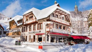 A traditional Alpine building with a snow-covered roof and wooden balconies, featuring a sign for a local restaurant and a view of a church spire in the background. - Hotel dasZimmermann (Todtmoos)
