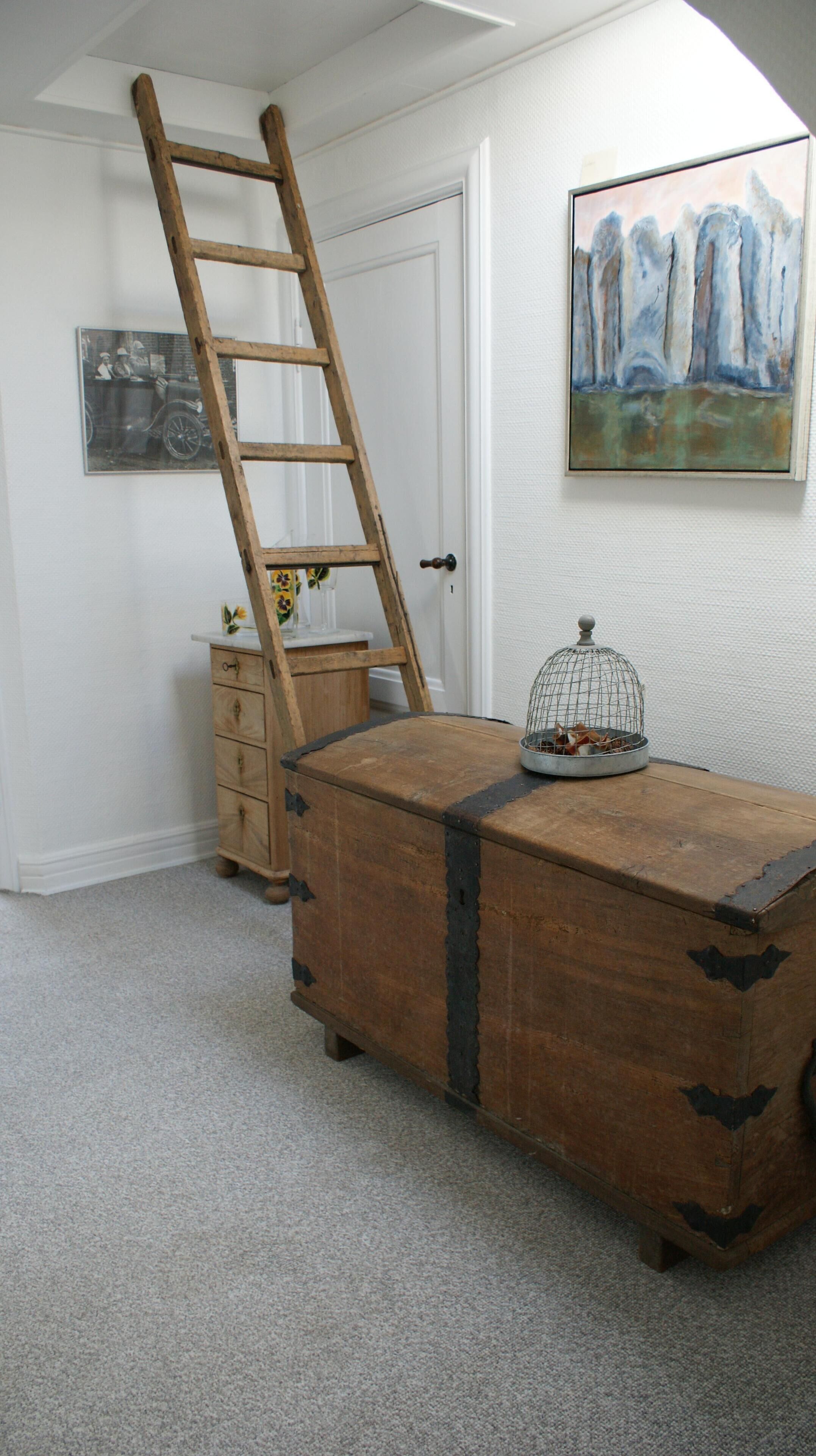 Een kamer met een houten kist, een ladder tegen een deur en een foto van een klassieke auto, plus een schilderij aan de muur.