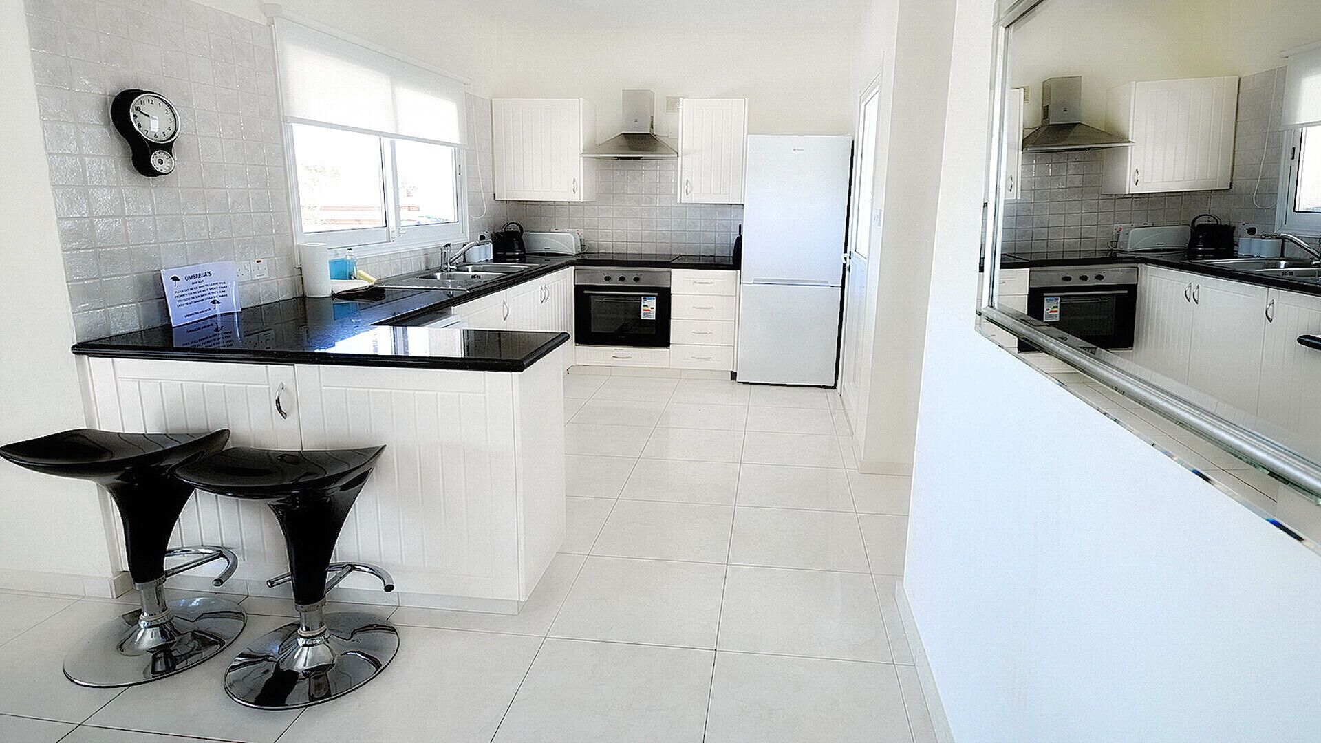 A modern kitchen with white cabinets, a black countertop, and a built-in oven. There are two bar stools with black seats.