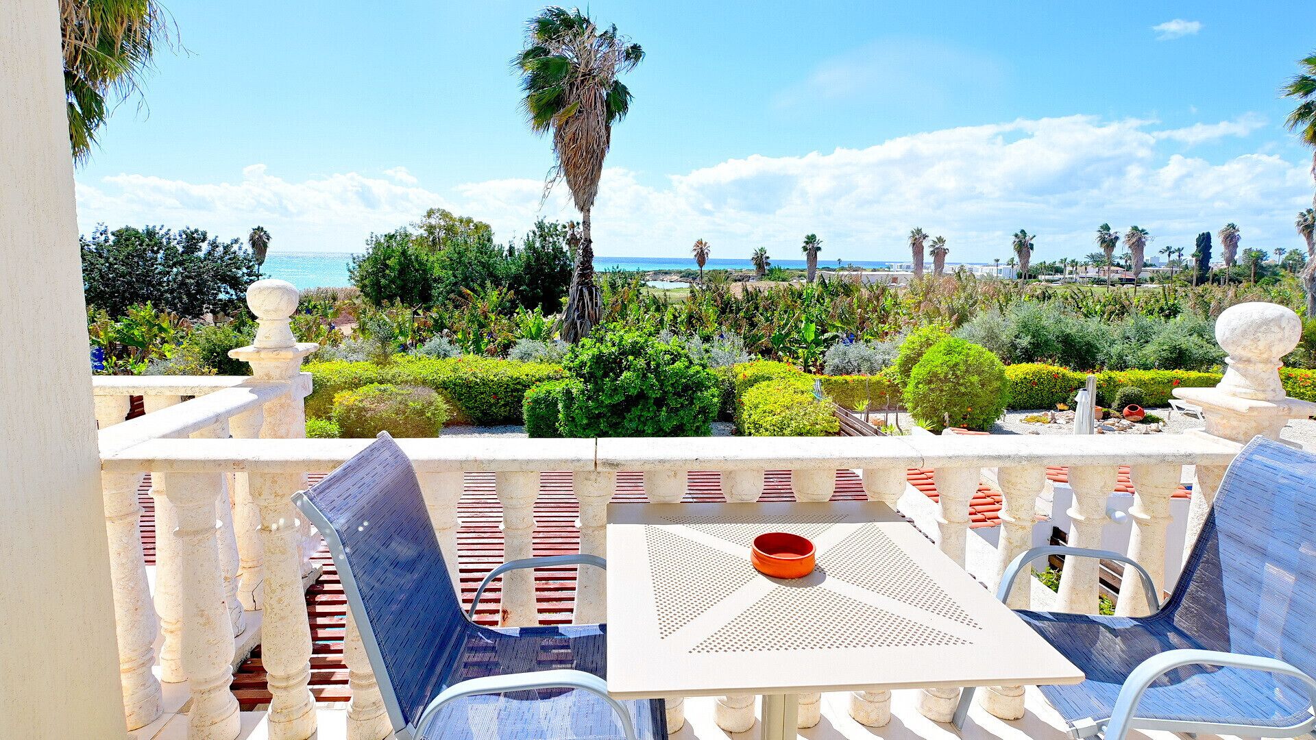 A balcony with a table and chairs, overlooking a landscaped garden and the sea.