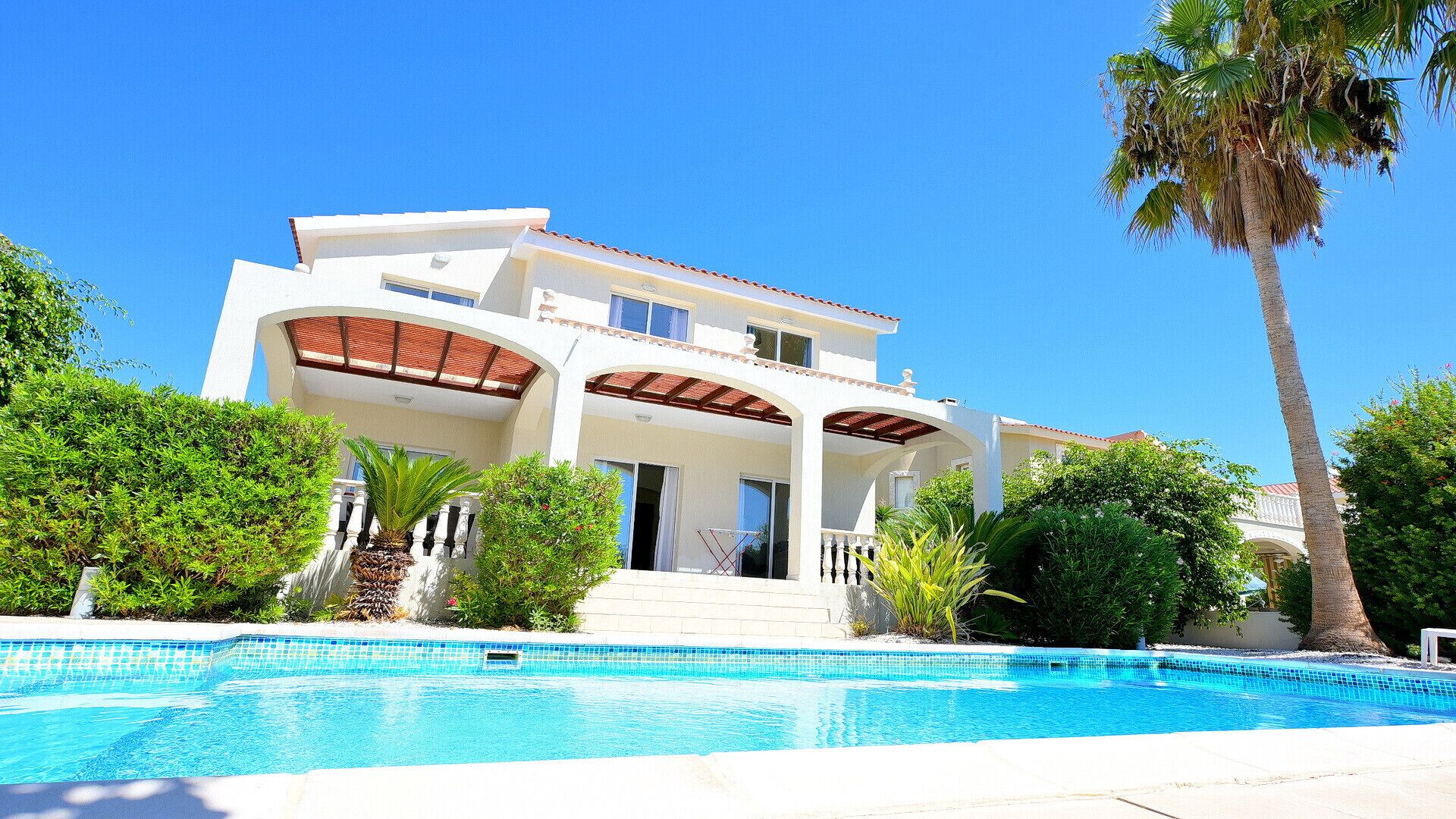 A two-story house with a swimming pool, surrounded by greenery and palm trees.