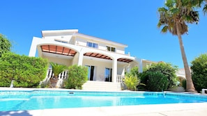 A two-story house with a swimming pool, surrounded by greenery and palm trees.
