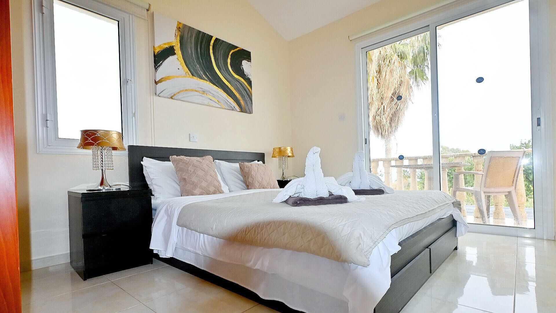 A bedroom with a large bed, two bedside tables, a painting on the wall, and a balcony with a view.