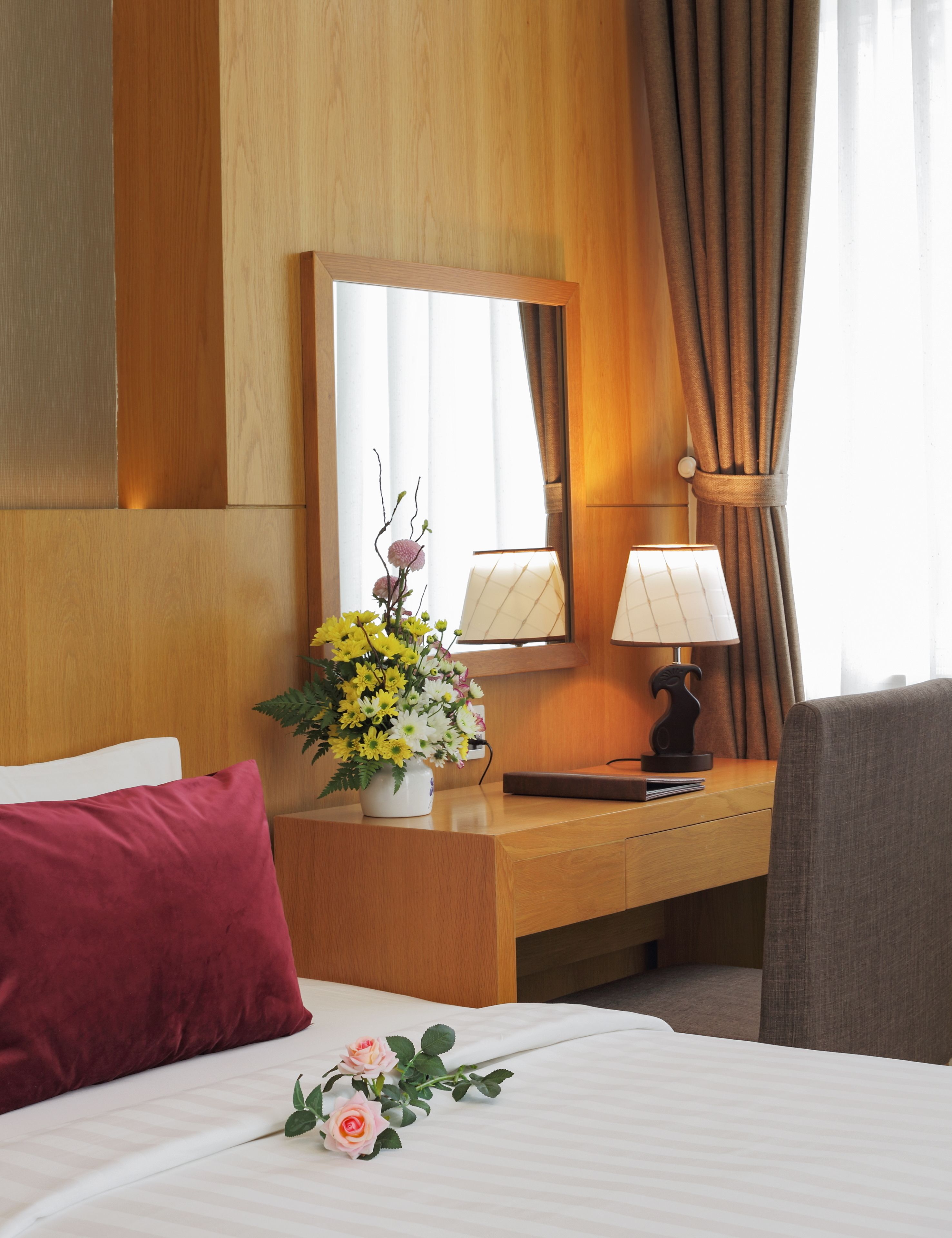 A hotel room with a bed, a wooden nightstand with a lamp, a mirror, and a vase of flowers.