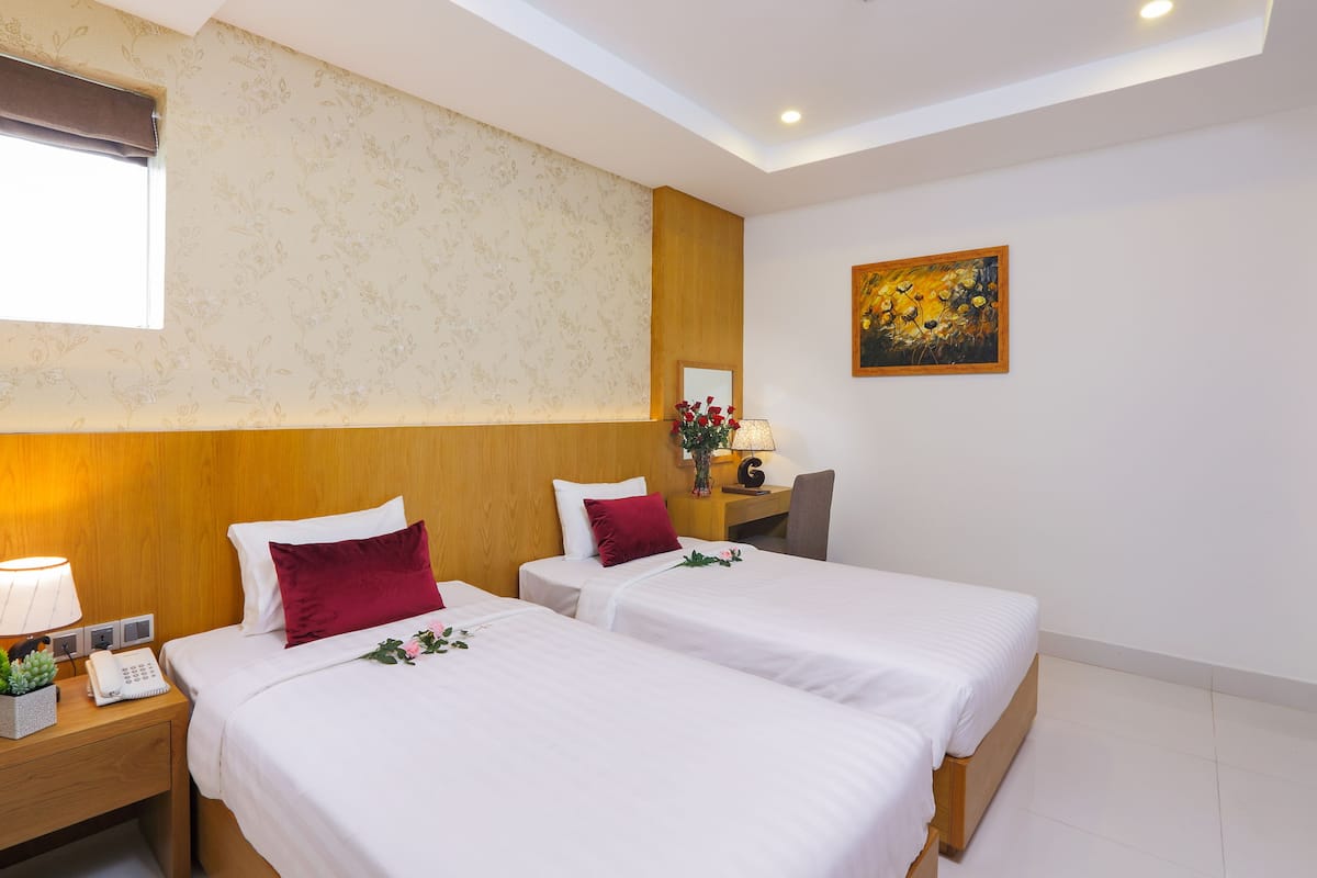 Une chambre d’hôtel avec deux lits, une tête de lit en bois, une table de chevet avec une lampe, une petite table avec un vase et un tableau accroché au mur.