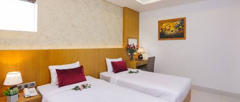 A hotel room with two beds, a wooden headboard, a nightstand with a lamp, a small table with a vase, and a painting on the wall.