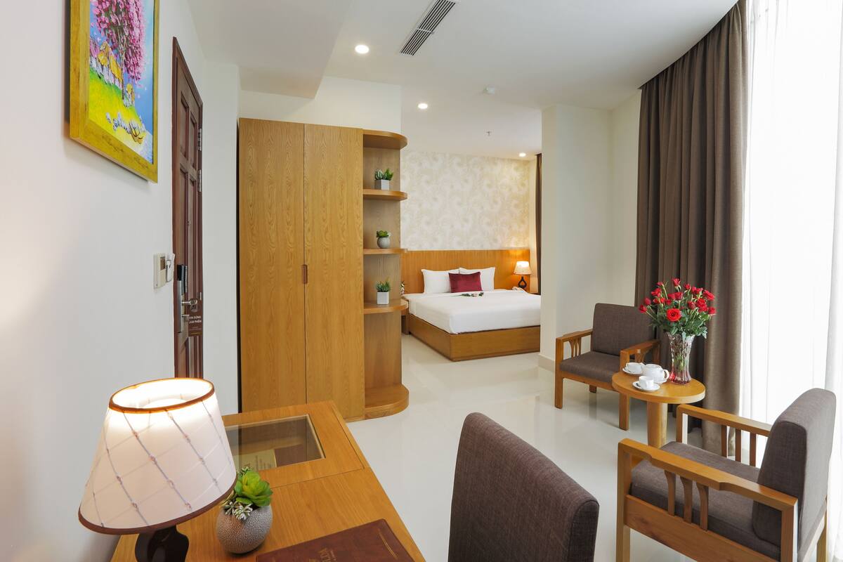 Une chambre d’hôtel avec un lit, un bureau doté d’une lampe, une chaise et un canapé. Il y a un tableau au mur et une fenêtre avec des rideaux.