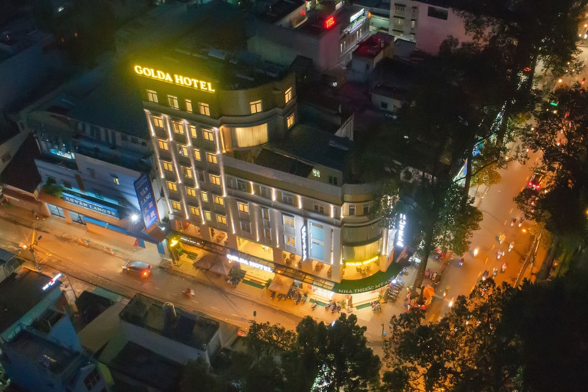 Un hôtel à plusieurs étages, reconnaissable à son grand panneau lumineux portant l’inscription « GOLD HOTEL », entouré d’autres immeubles et bordé par une rue animée.