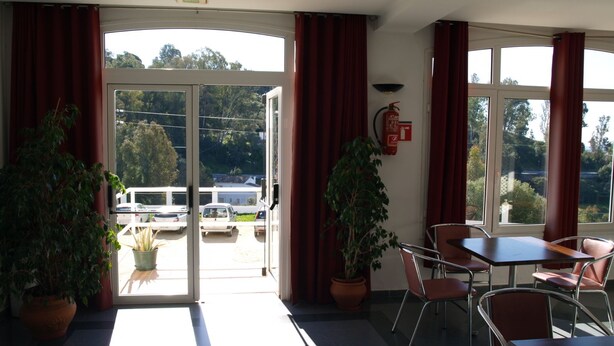Un hall d’hôtel donnant sur un parking et sur des arbres à l’extérieur. On y trouve des plantes en pot, un extincteur et une table avec des chaises.