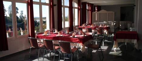 A dining area with multiple tables set for a meal, large windows with curtains, and a view of the outdoors.
