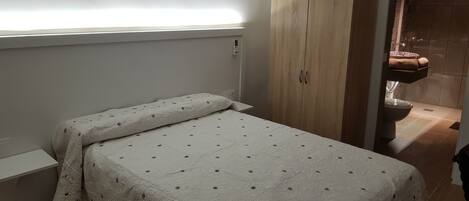 Una habitación pequeña con cama individual, un armario de madera y un baño visible a través de una puerta abierta.