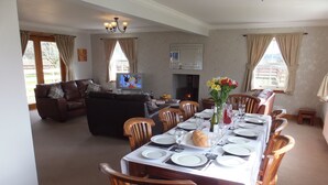A dining area with a long table set for six, surrounded by wooden chairs. A television is mounted on the wall, and a fireplace is visible in the background. - A Beautiful Rural Farmhouse With Flexible Living Space And Great Views (Stirling)