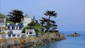 A coastal building with a tower, surrounded by trees and a rocky shoreline. - Feet in the water, wooden house with terrace, breathtaking sea view. (Locquirec)