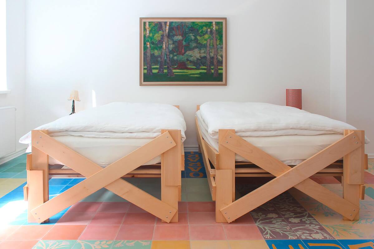 Two wooden bunk beds with white bedding, a painting of a forest on the wall, and a colorful tiled floor.
