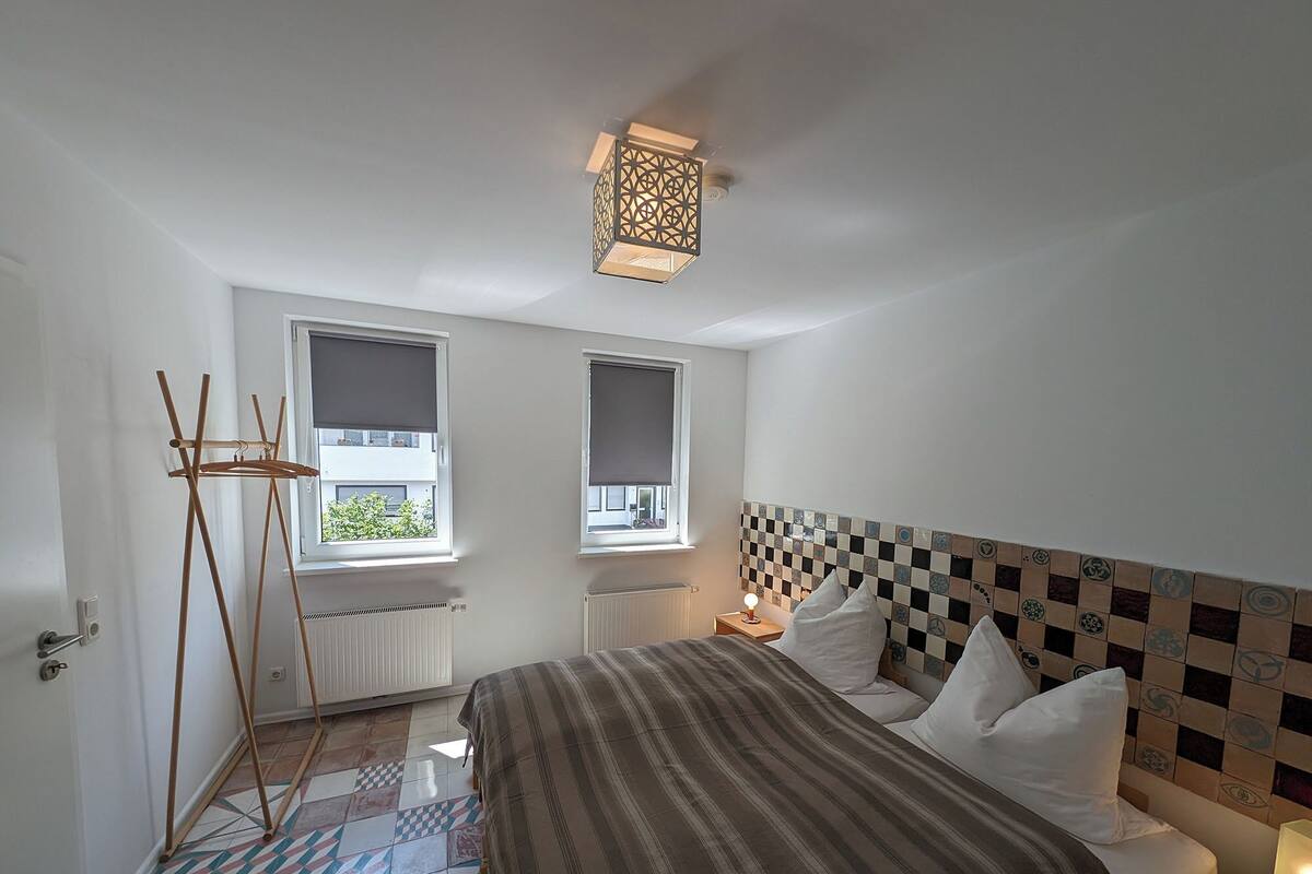 A bedroom with a bed, a radiator, a wooden drying rack, two windows with blinds, and a patterned tile floor.