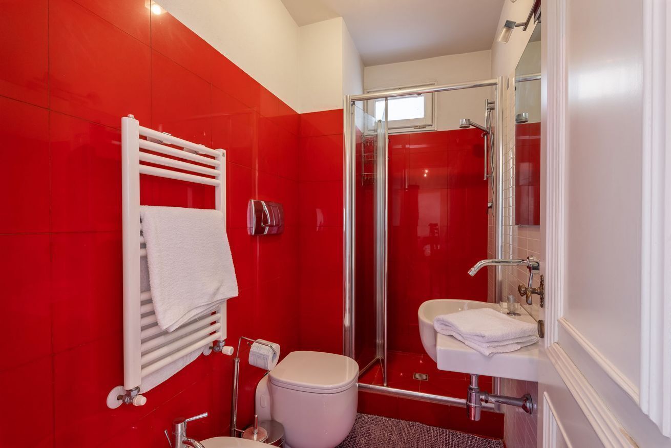 Une salle de bain avec un mur recouvert de carreaux rouges, des toilettes blanches, une douche en verre et un lavabo blanc.