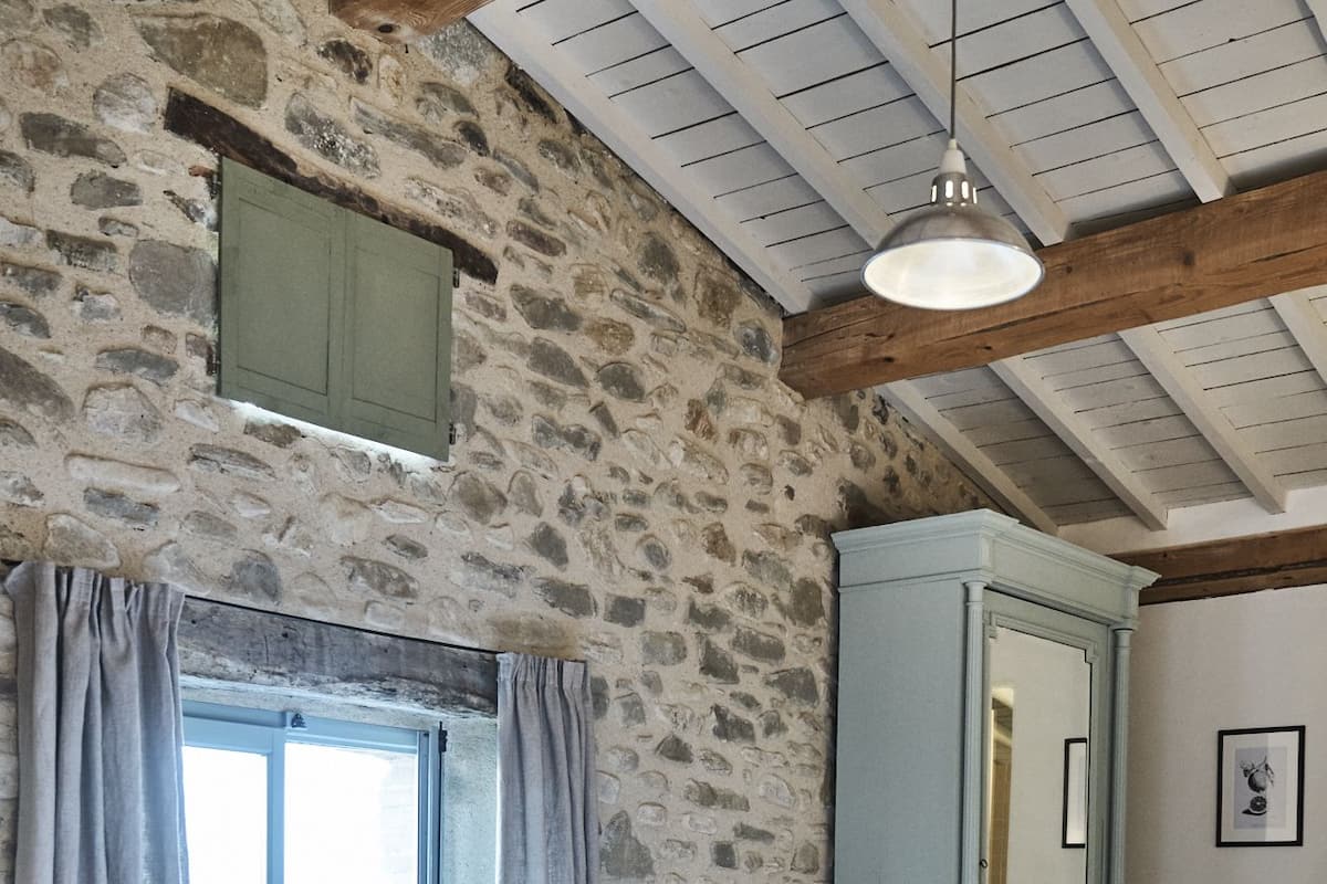 A bedroom with stone walls, a wooden ceiling, a bed, a wardrobe, and a window with curtains.