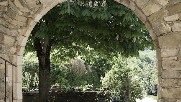 Une structure en pierre en forme d’arche, avec un chemin de gravier qui traverse un jardin agrémenté de plantes en pot et d’arbres.