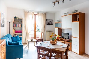 A well-lit living room with a blue sofa, a dining table with chairs, a bookshelf, a TV, and a cabinet.