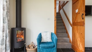 Een kamer met een zwart houtkacheltje, een blauwe fauteuil, een rieten mand en een houten trap.