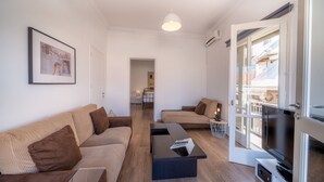A modern living room with two sofas, a coffee table, and a TV. There is a door leading to a balcony with a view of buildings. - The Roof - Flat Sea View in Aegina Town (Aegina)