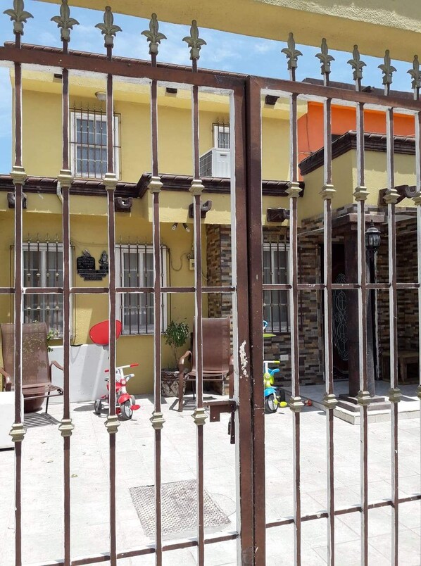 Edificio amarillo con entrada protegida por un portón, patio con baldosas y algunos sillones y juguetes fuera.