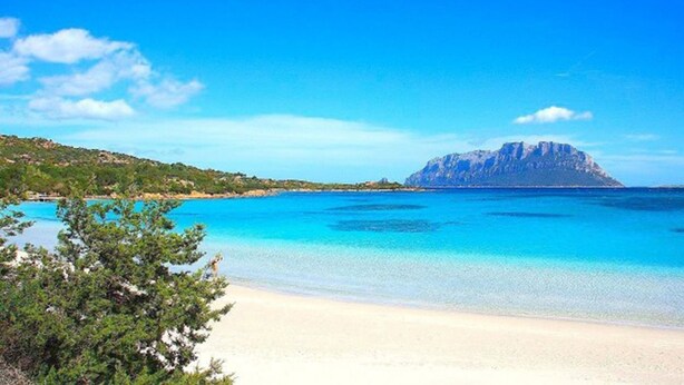 Une plage aux eaux turquoise et limpides, un rivage sablonneux et une montagne en arrière-plan.