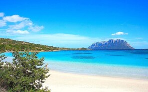 Ein Strand mit klarem türkisblauem Wasser, einem sandigen Ufer und einem Berg im Hintergrund.