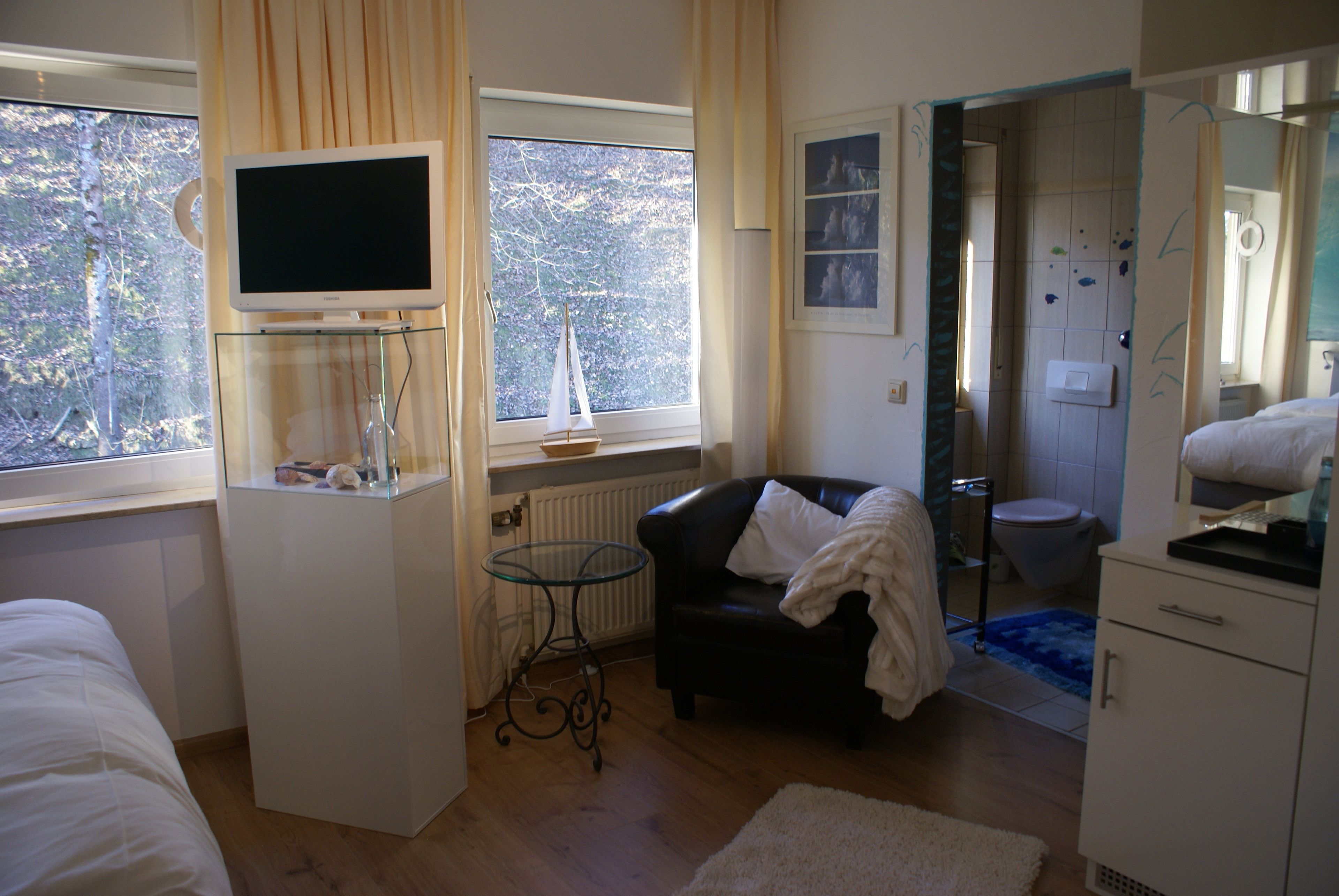 A hotel room with a bed, a desk with a computer, a chair, a window with a view of trees, and a bathroom with a toilet and sink.