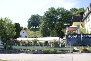 Um hotel com uma placa que diz "Hotel - Restaurante" e um mastro com uma bandeira. Há árvores e uma colina ao fundo.
