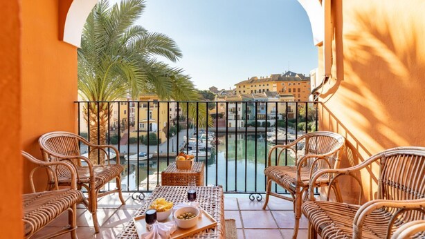 A balcony with wicker furniture, a table with fruits and drinks, and a view of a canal with buildings in the background. - A (VA022) TH Casa Port Saplaya (Alboraya)