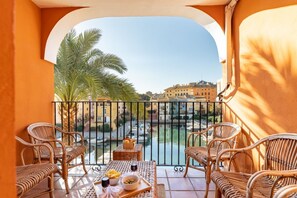 Un balcone con mobili in vimini, un tavolo con frutta e bevande, e vista su un canale con edifici sullo sfondo.