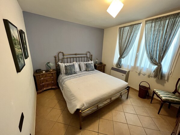A bedroom with a bed, bedside tables, a nightstand, a chair, a radiator, and curtains.