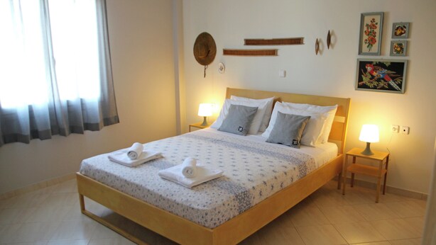 A neatly made bed with a floral bedspread, two pillows, and two rolled towels. There are two bedside lamps on wooden nightstands. The room has a tiled floor and a window with curtains. - Bellissima Campagna Home<br>Pelekapina Chania Crete  (Chania , Crete)