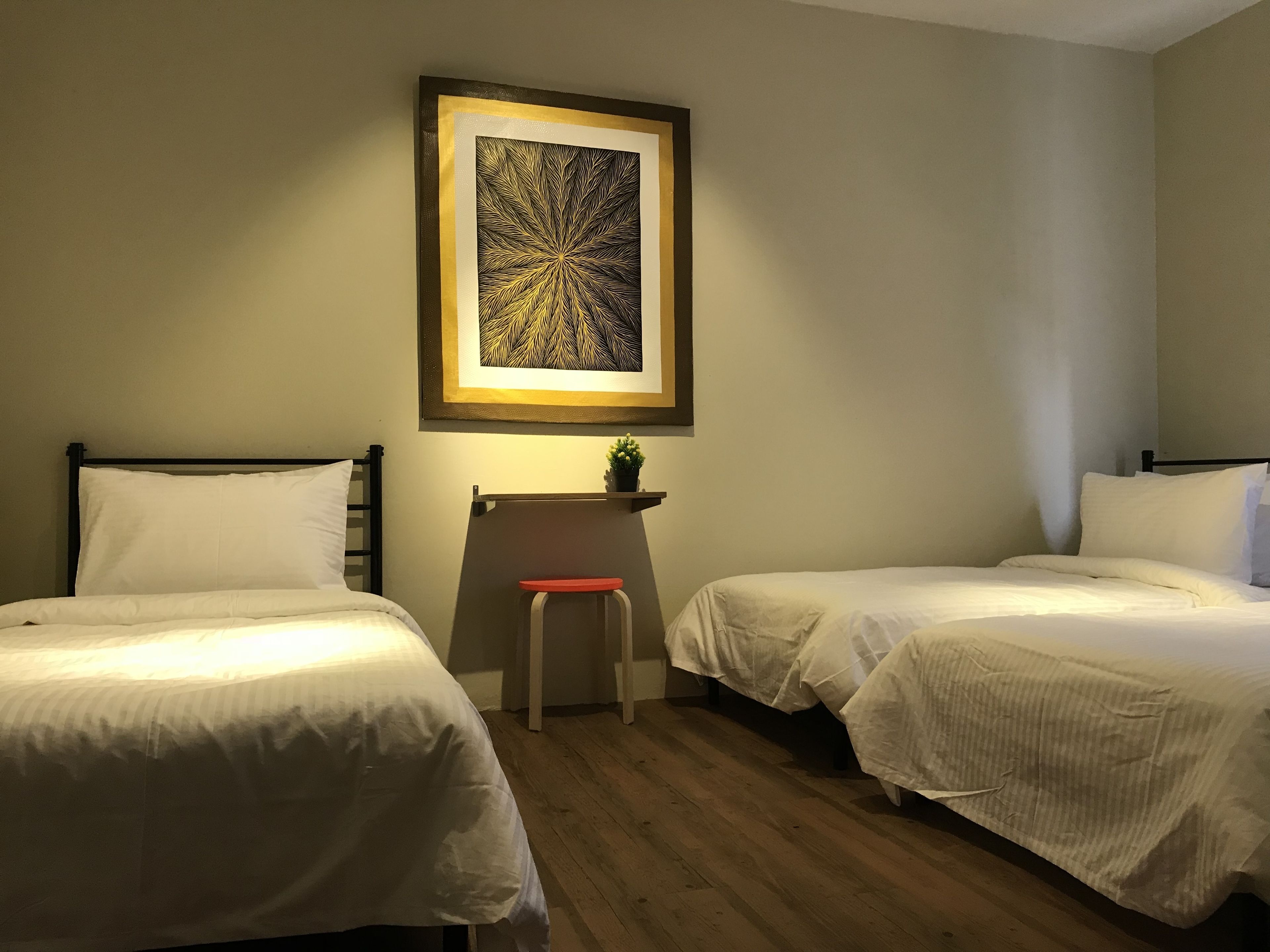 A hotel room with two single beds, a small stool, a framed artwork on the wall, and a shelf with a plant.