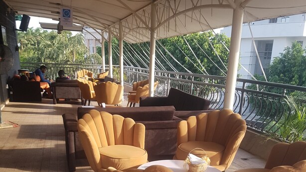 A covered outdoor seating area with yellow chairs and sofas, a tiled floor, and a railing overlooking greenery. - Zeina Suites Abuja (Abuja)