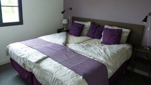 A neatly made bed with a purple and white bedspread, a bedside table with a lamp, and a window with a view of greenery.