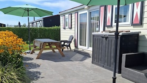 A patio area with a wooden table and chairs, a green umbrella, and a garden with yellow flowers.
