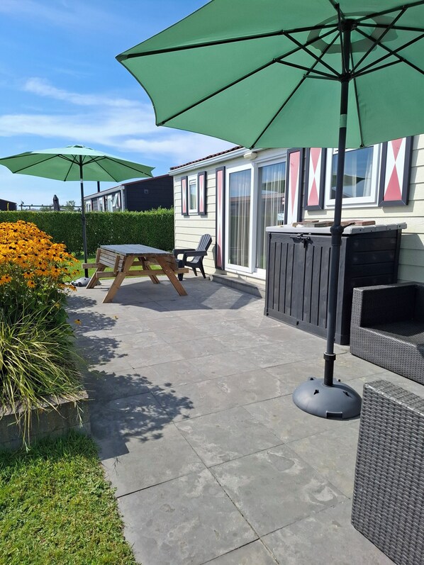 A patio area with a wooden table and chairs, a green umbrella, and a garden with yellow flowers.