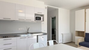 A modern kitchen with white cabinets, a microwave, and a sink. There is a dining table with chairs, a radiator, and a blue sofa in the adjacent living area. - Agi Rescator 218 Apartamento for rent in Roses Girona. (Roses)