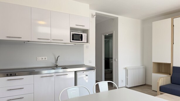 A modern kitchen with white cabinets, a microwave, and a sink. There is a dining table with chairs, a radiator, and a blue sofa in the adjacent living area. - Agi Rescator 218 Apartamento for rent in Roses Girona. (Roses)