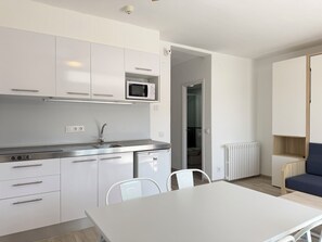 A modern kitchen with white cabinets, a microwave, and a sink. There is a dining table with chairs, a radiator, and a blue sofa in the adjacent living area. - Agi Rescator 218 Apartamento for rent in Roses Girona. (Roses)