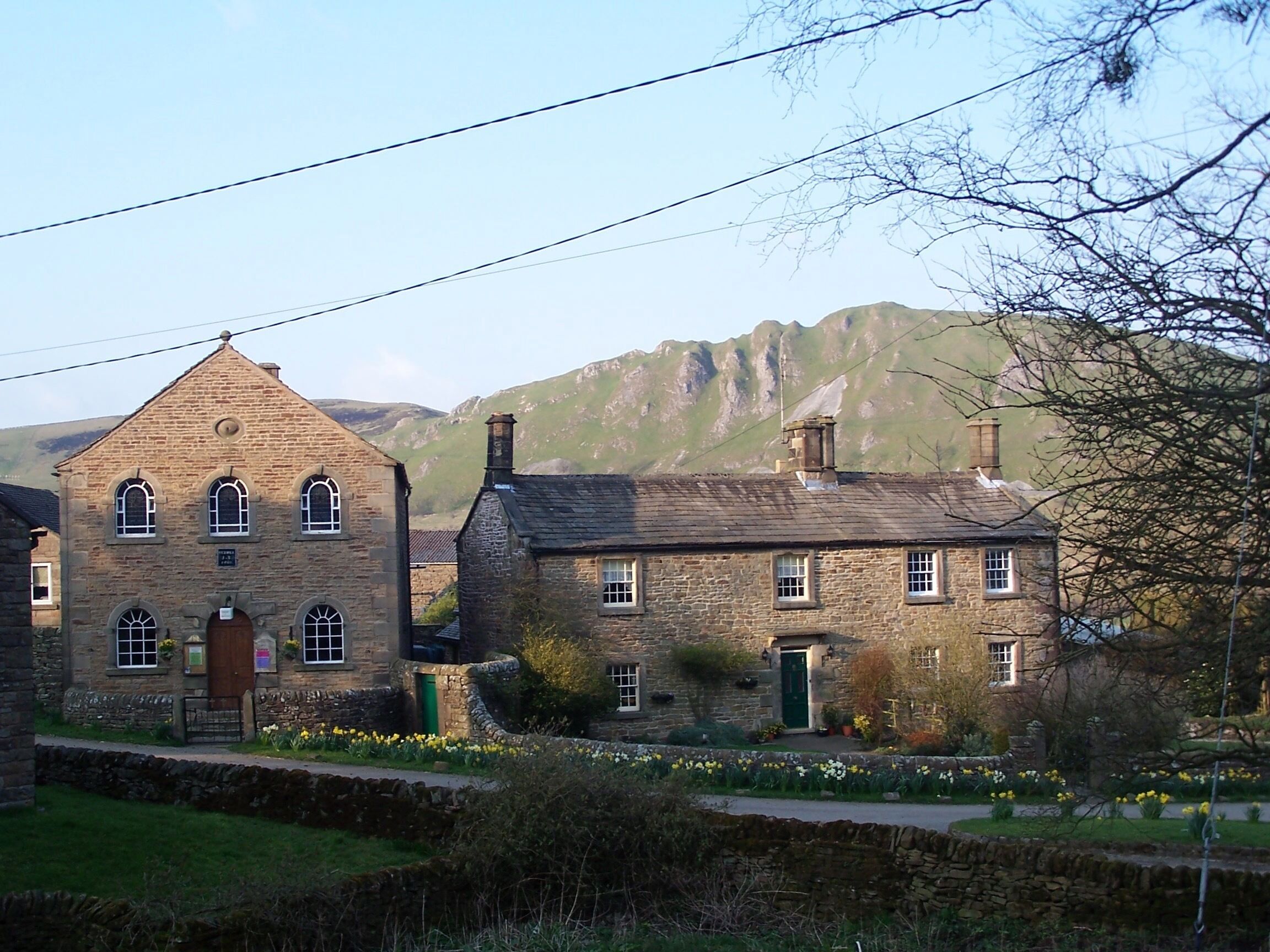 A stone building with a triangular roof and multiple windows, surrounded by a stone wall and a garden with daffodils.