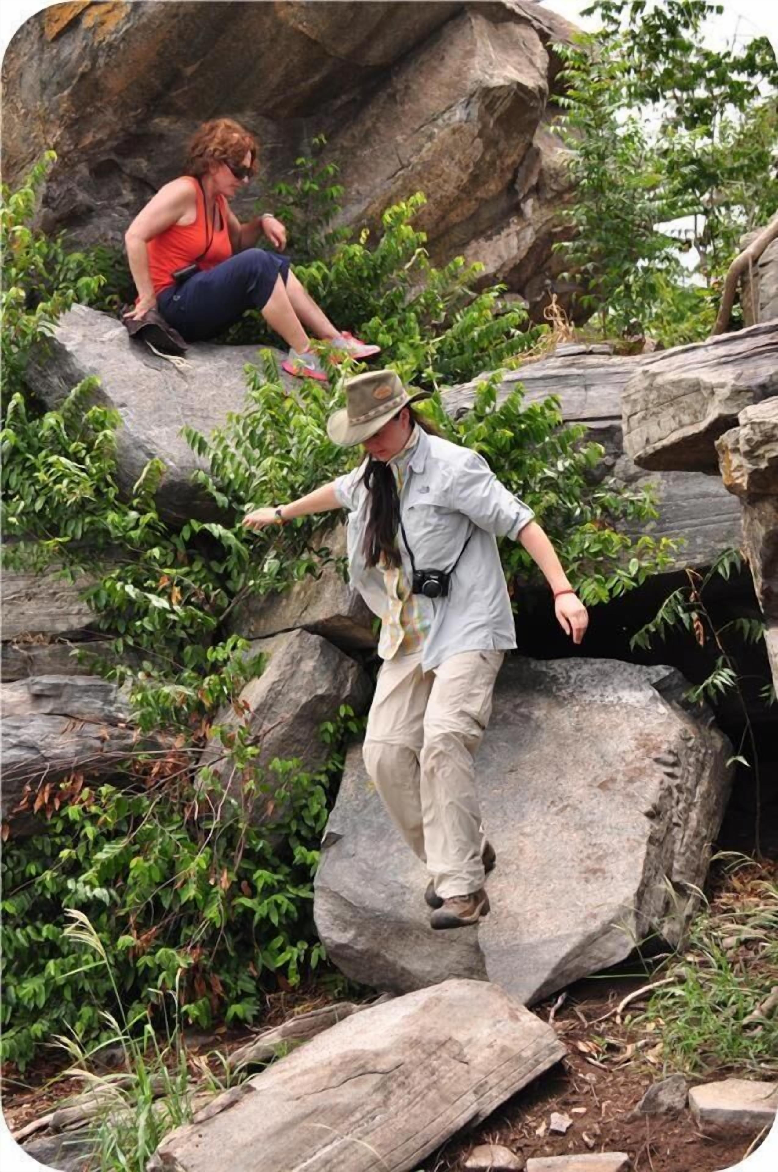 Two people sitting on large rocks in a natural setting with greenery around.