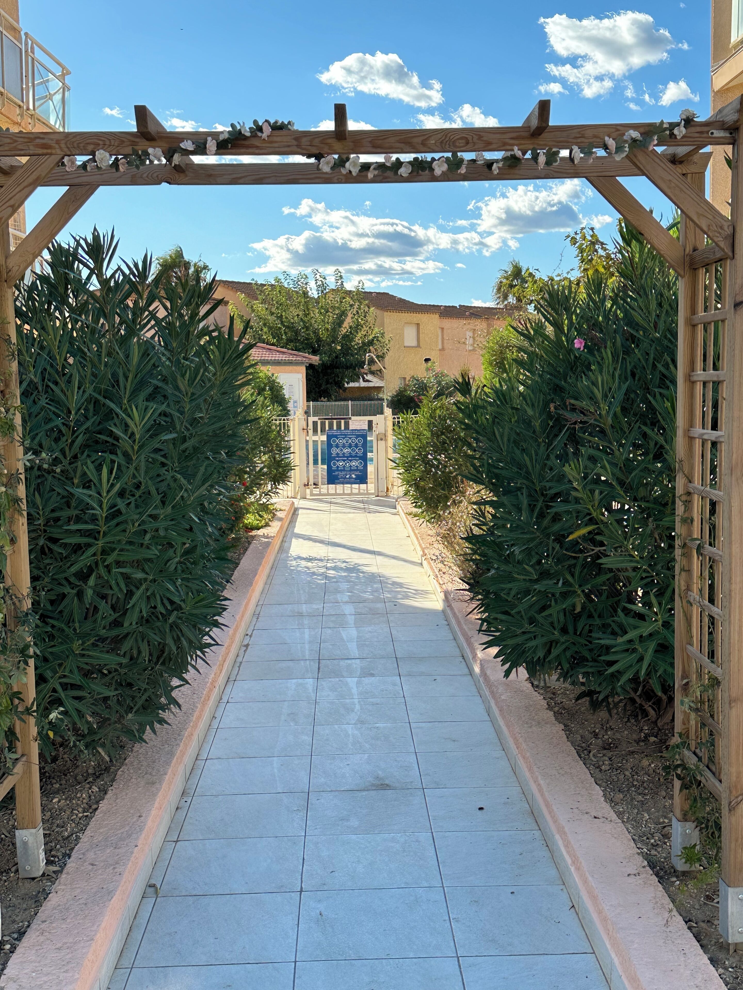 A pathway leading to a gated entrance with a blue sign, flanked by green plants and a wooden pergola.