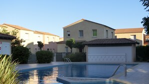 Una piscina amb vora de rajola, envoltada per un passeig pavimentat. S'hi veuen edificis al fons amb teulada de rajola i una porta de garatge.